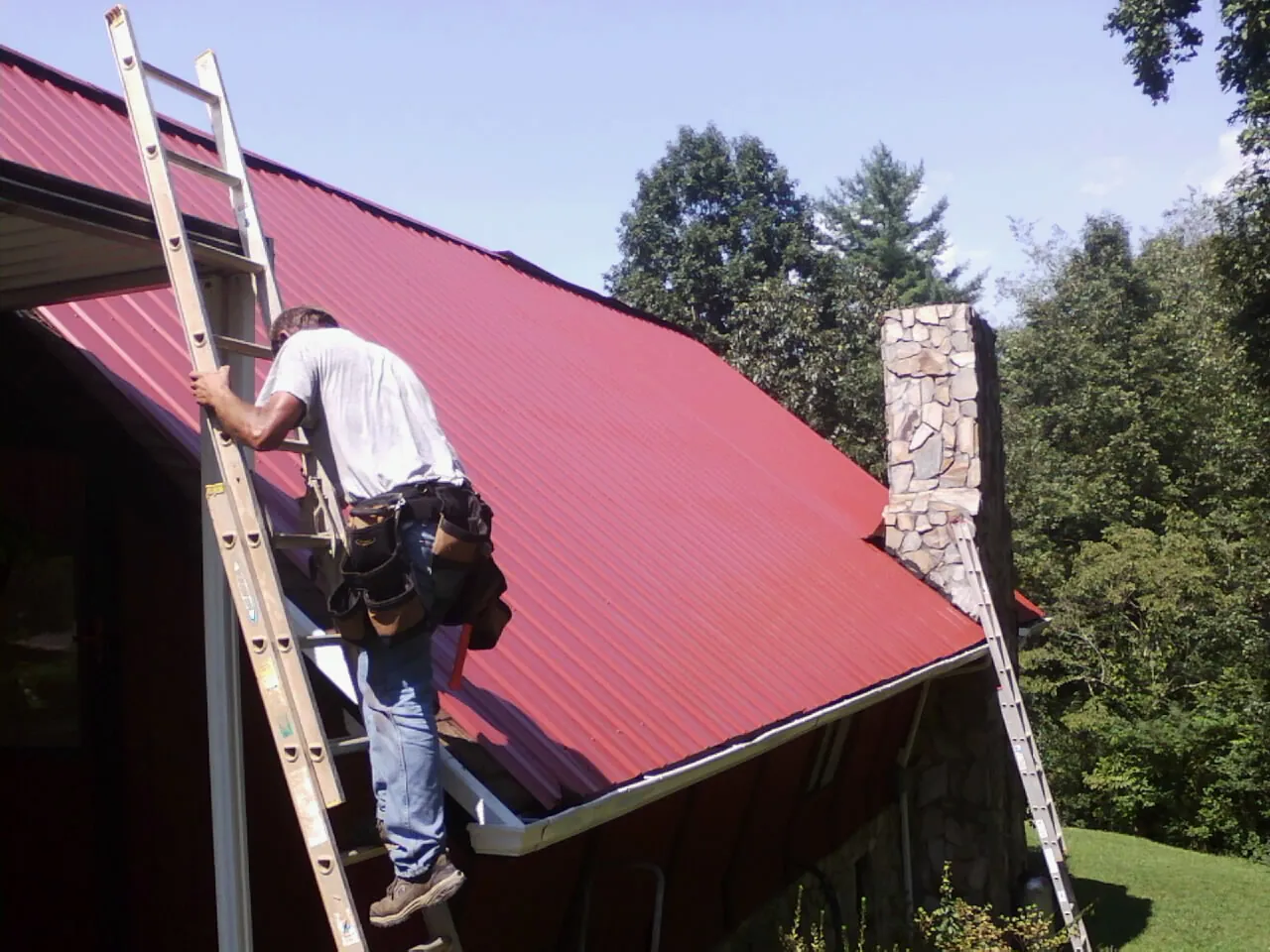 Roofer on a ladder beside a red metal roof and stone chimney — Soffit & Fascia Repair by Roofing Brothers in Tuscarora